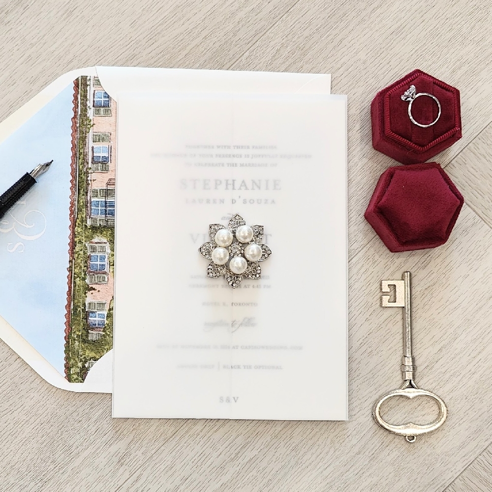 Single card wedding invite with a vellum wrap and pearl brooch embellishment.  It is paired with an envelope with a venue sketch inspired envelope liner.