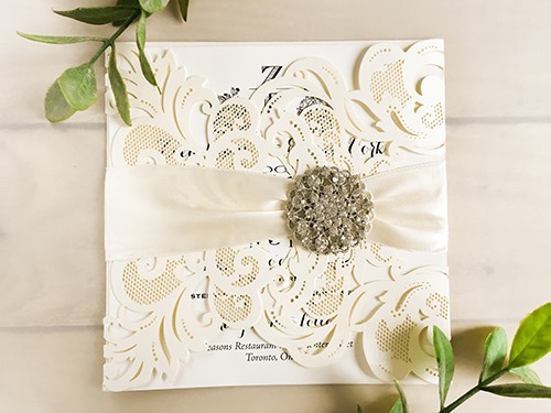 Invitation lc20: Ivory Shimmer, Cream Smooth, Antique Ribbon, Brooch/Buckle X - This is a laser cut wedding invitation in a gate fold design.  The paper is the ivory shimmer.  There is an antique ribbon and brooch on the front.